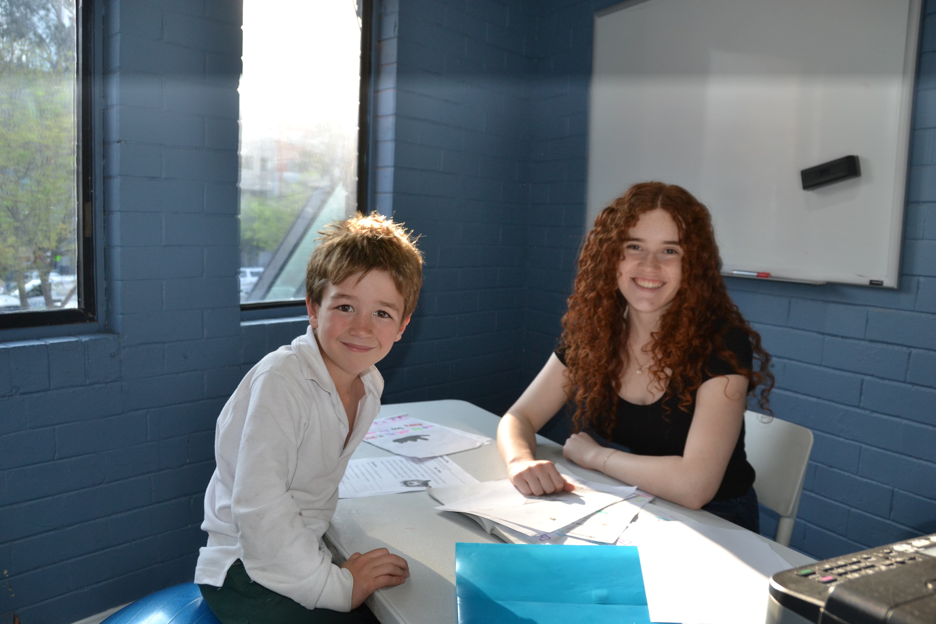 A First Education tutor and a student sitting together at a centre desk with worksheets, near a whiteboard and window.