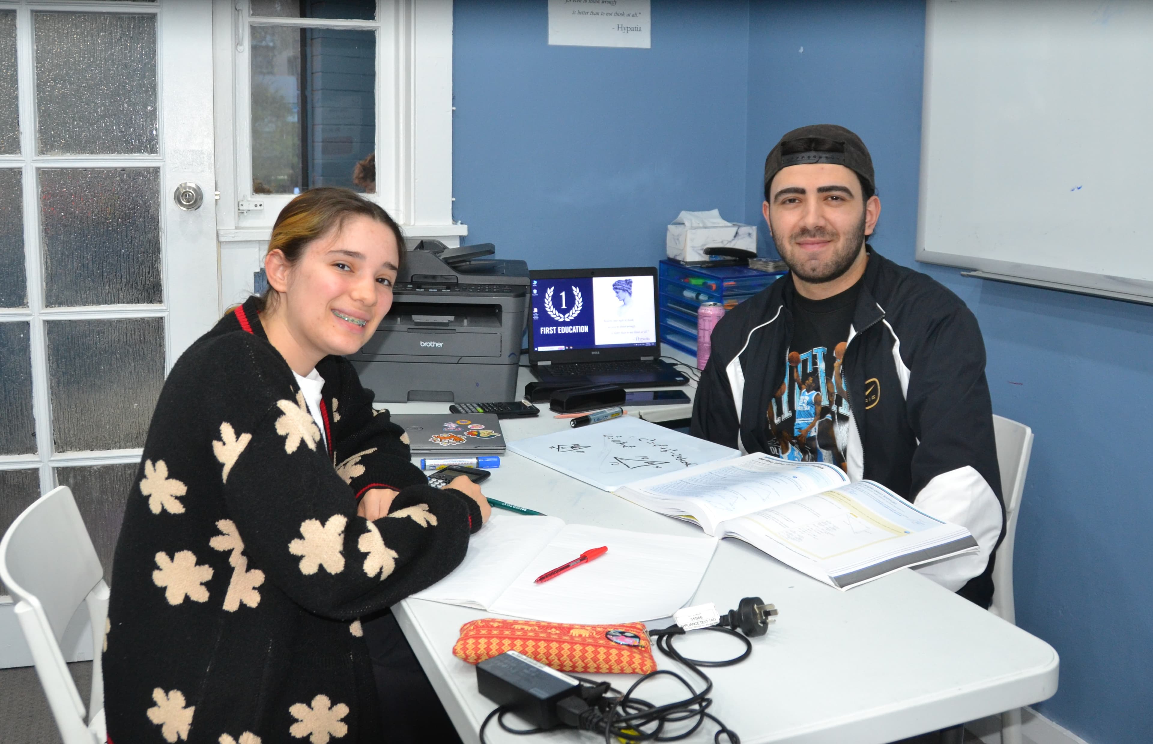 A First Education tutor and a student giving thumbs up at a centre desk with open workbooks and a tablet.