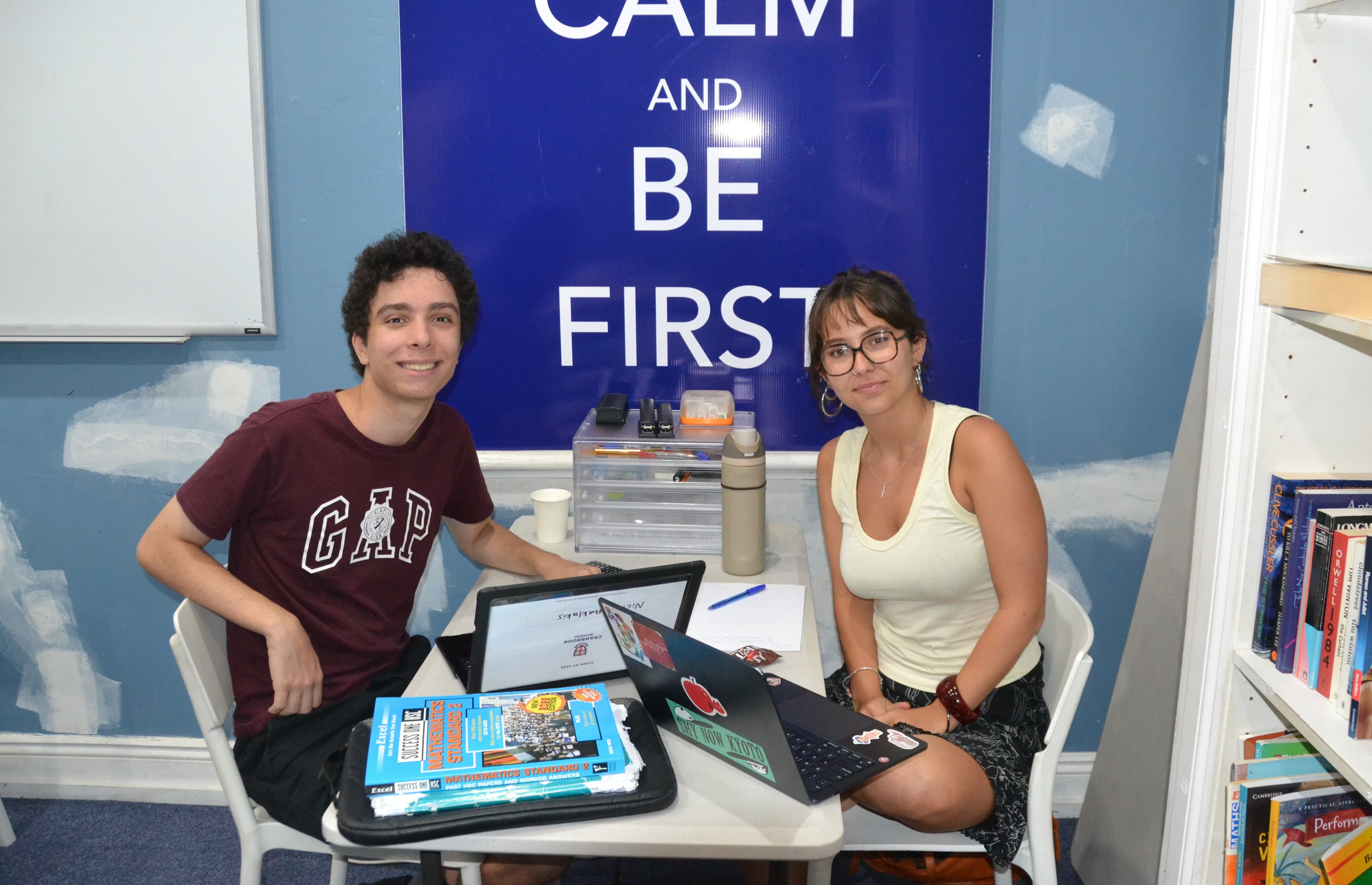 A First Education tutor and a student sitting together at a centre desk with laptops and HSC study guides, in front of a bookshelf.