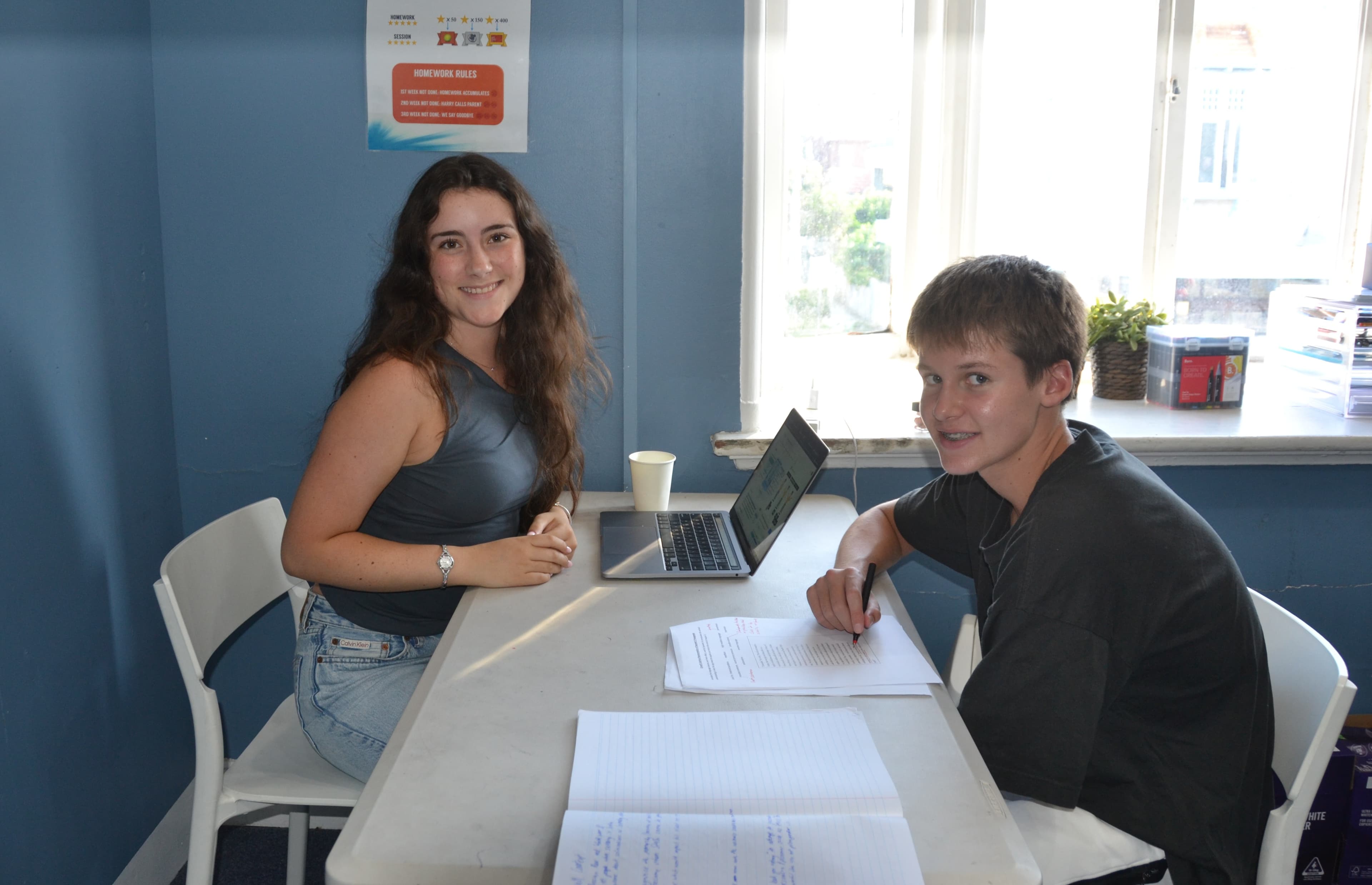 A First Education tutor and a student sitting together at a centre desk with an open laptop and handwritten notes, near a window.