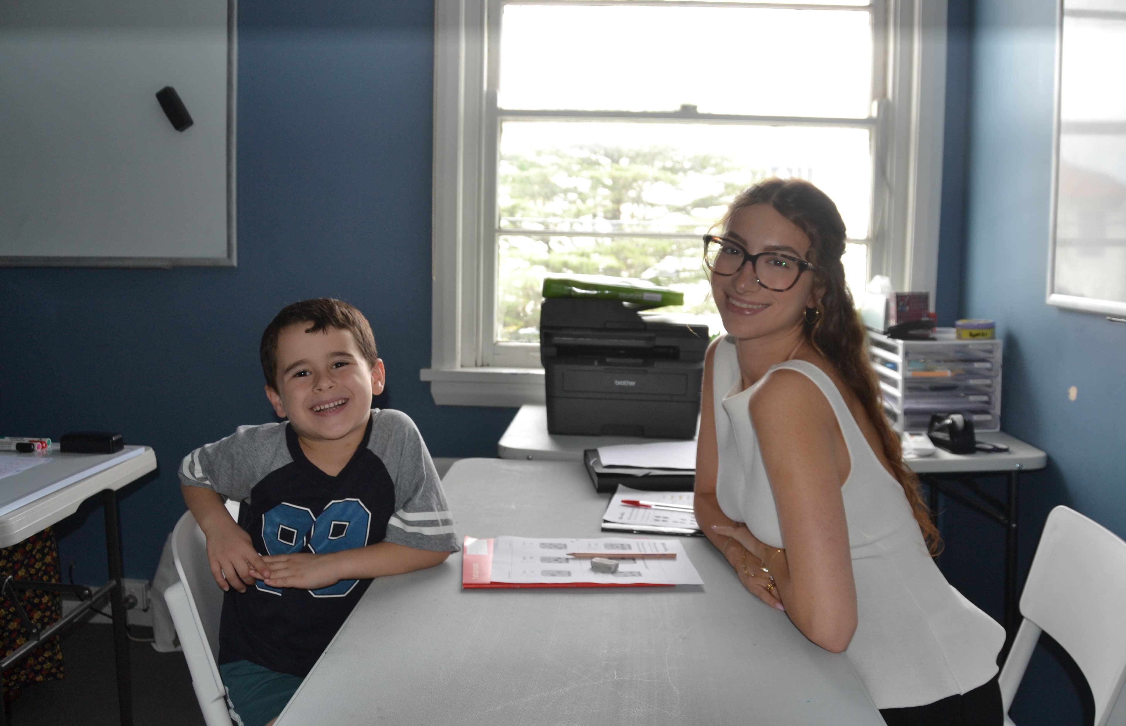 A First Education tutor and a kindergarten student sitting together at a centre desk with worksheets and study materials.