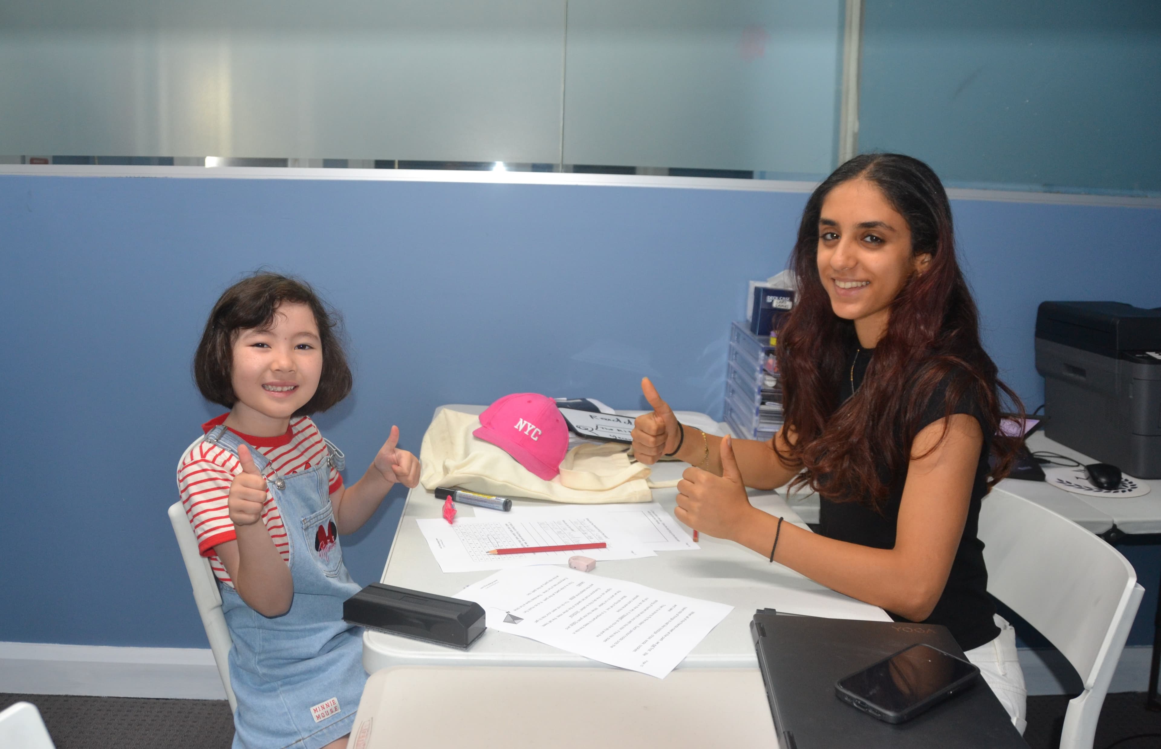 A First Education tutor and a kindergarten student sitting together at a centre desk with open workbooks and study papers.