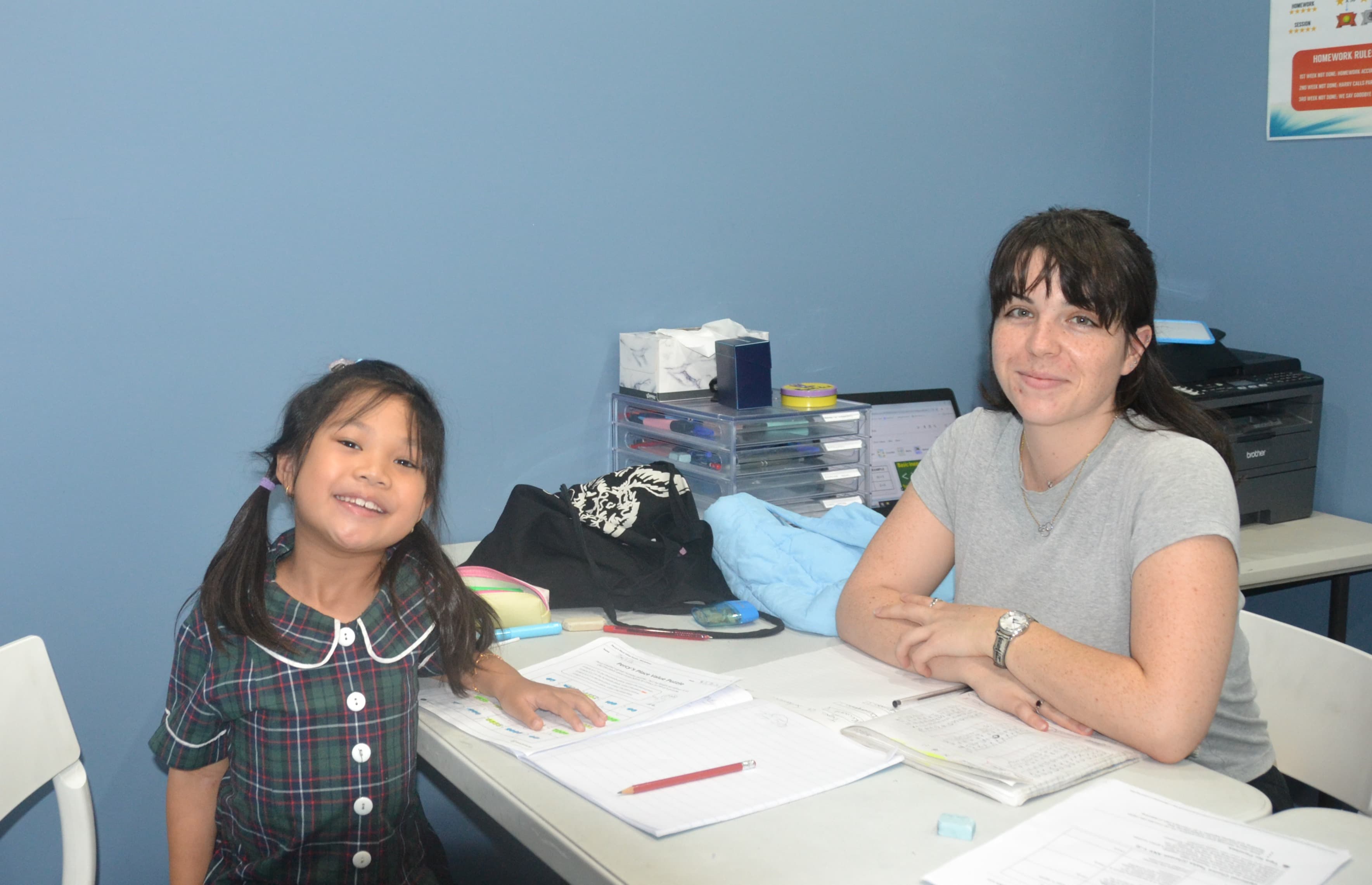A First Education tutor and a kindergarten student giving double thumbs up at a centre desk with worksheets and study materials.