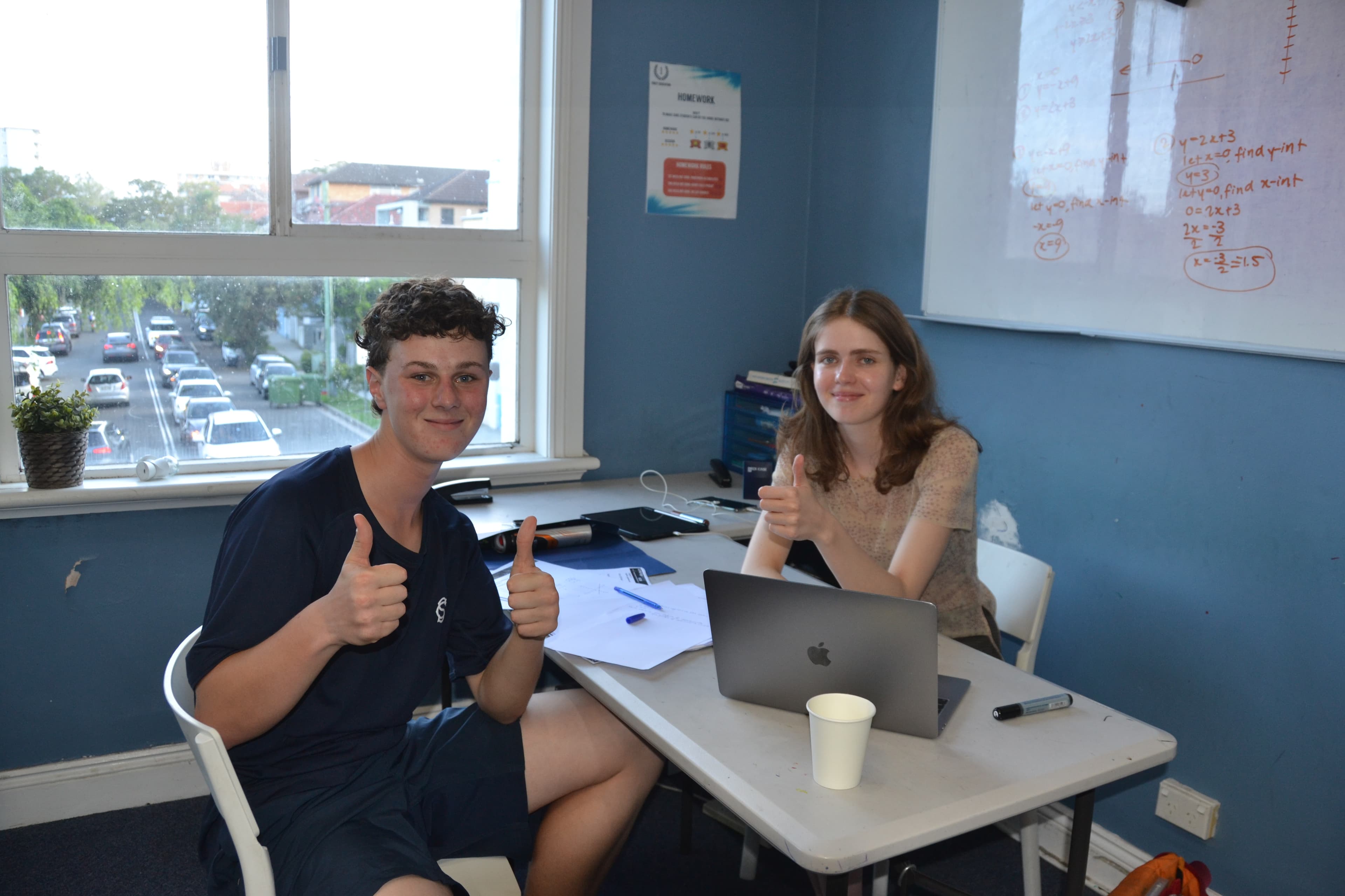 A First Education tutor and a student giving double thumbs up at a centre desk with an open laptop, in front of a whiteboard covered in maths workings.