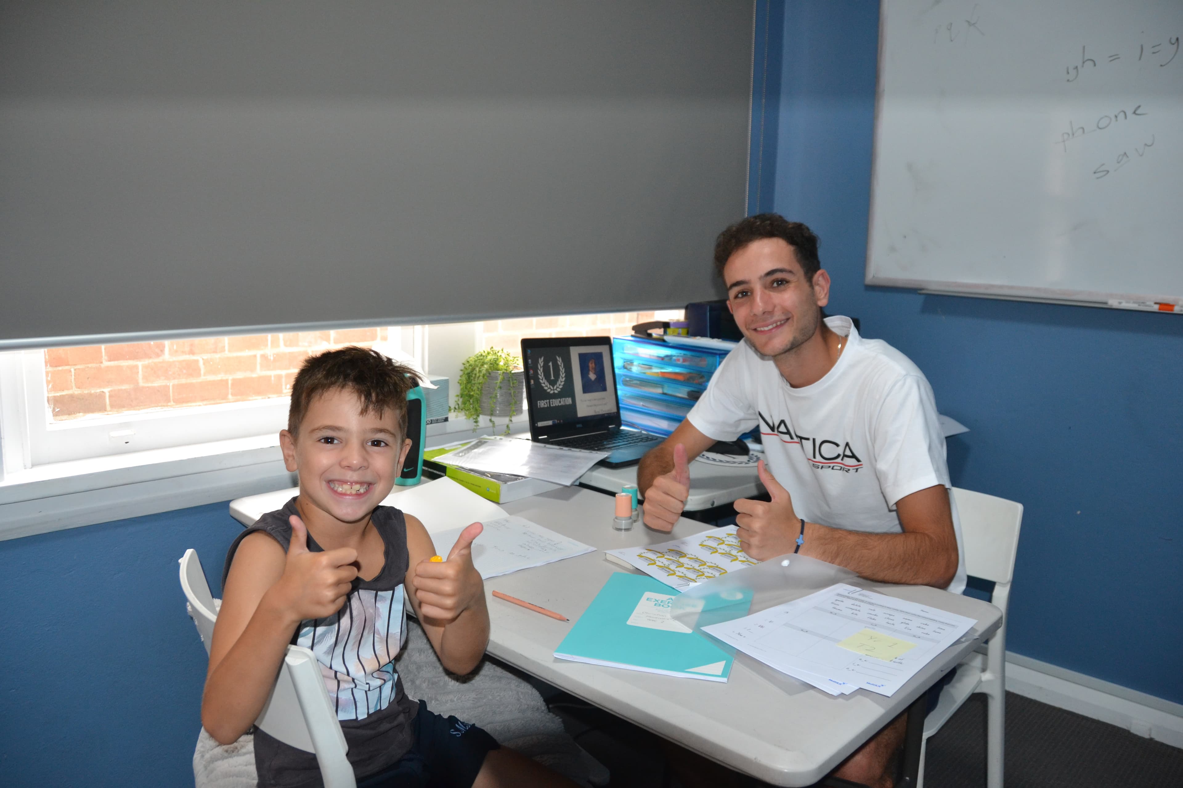 A First Education tutor and a Year 1 student giving double thumbs up at a centre desk with open workbooks.