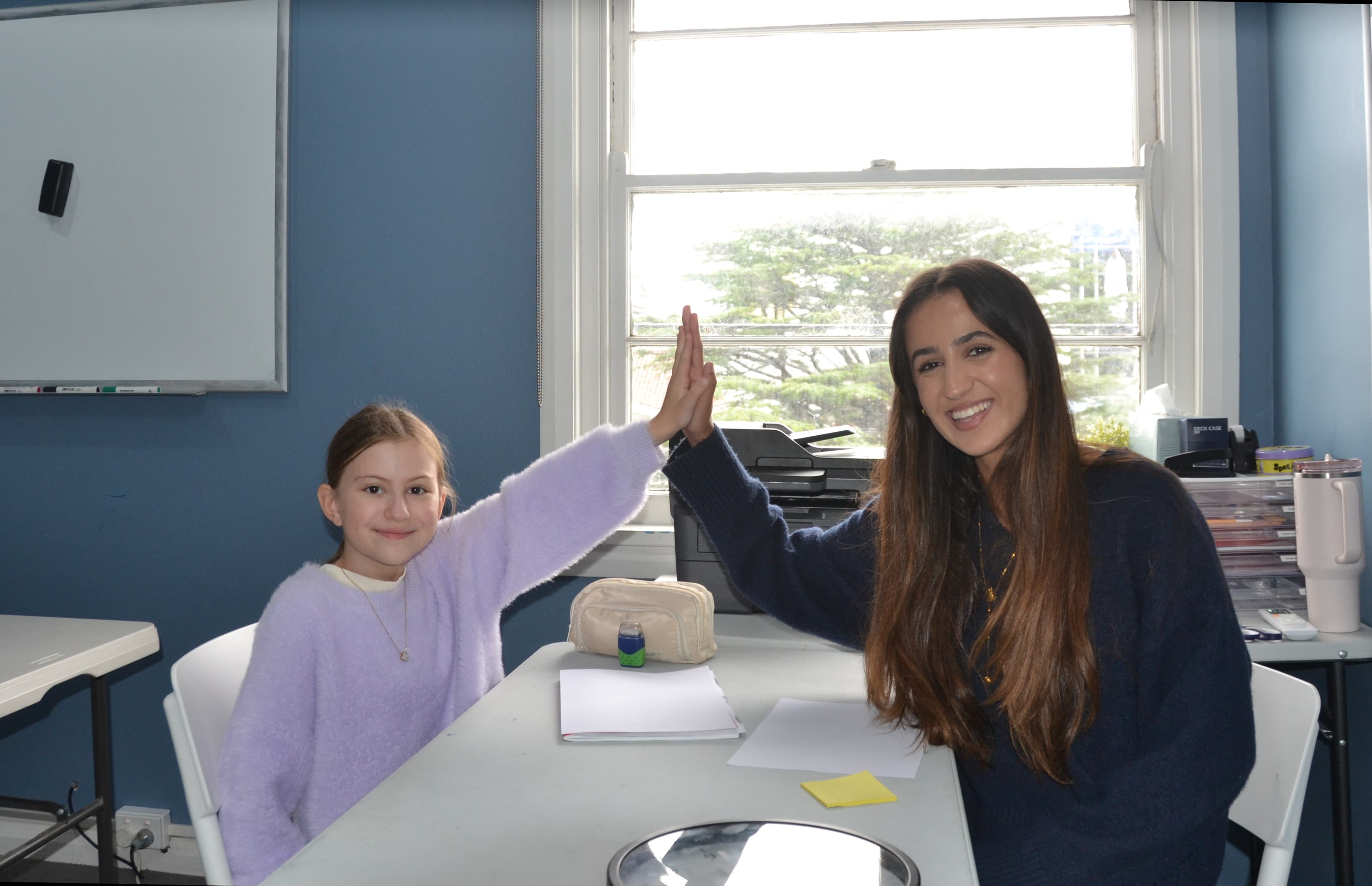 A First Education tutor and a Year 1 student giving a high five at a centre desk with worksheets and study notes.