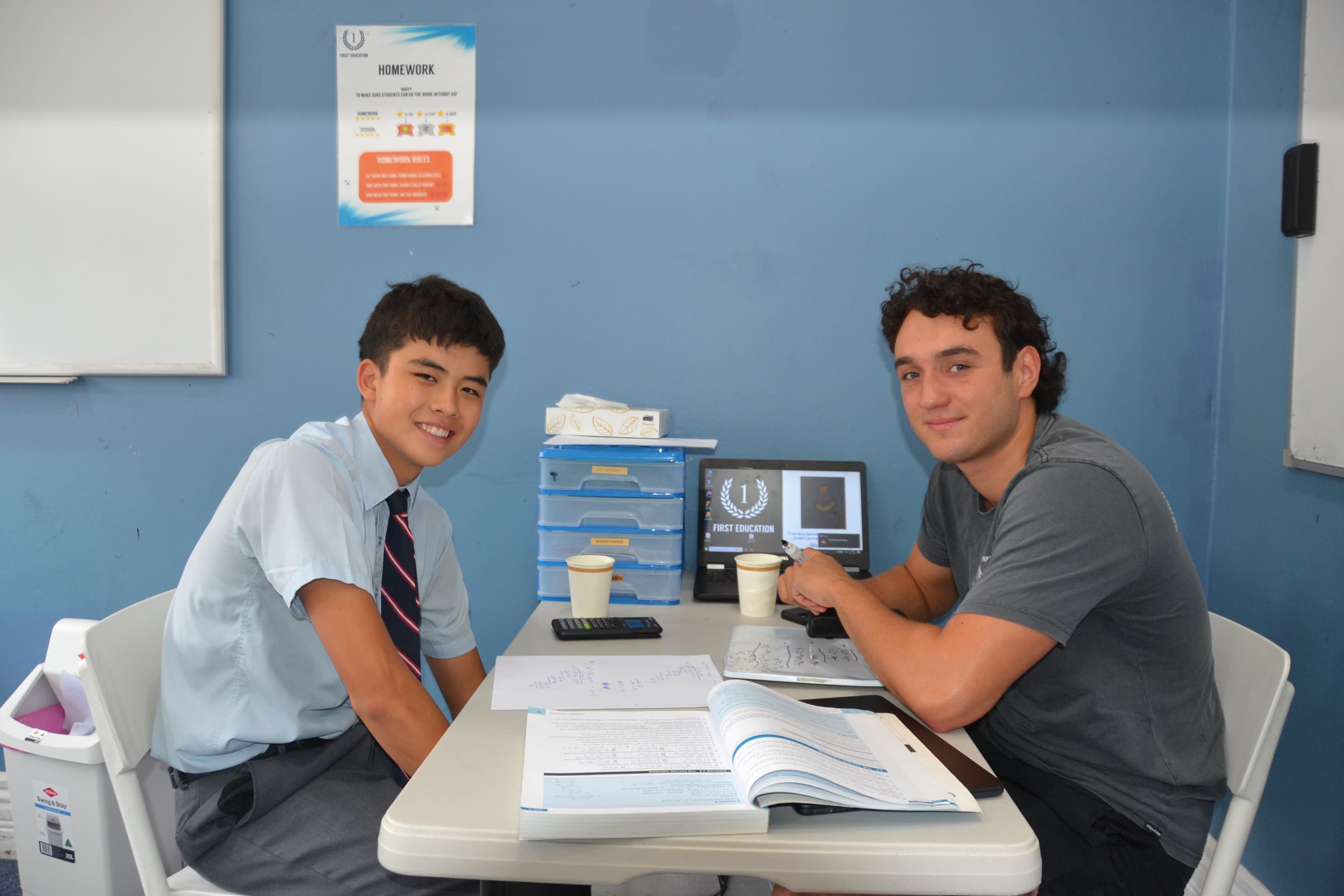 A First Education tutor sitting with a Year 10 student in school uniform, working through a maths textbook together at a centre desk.