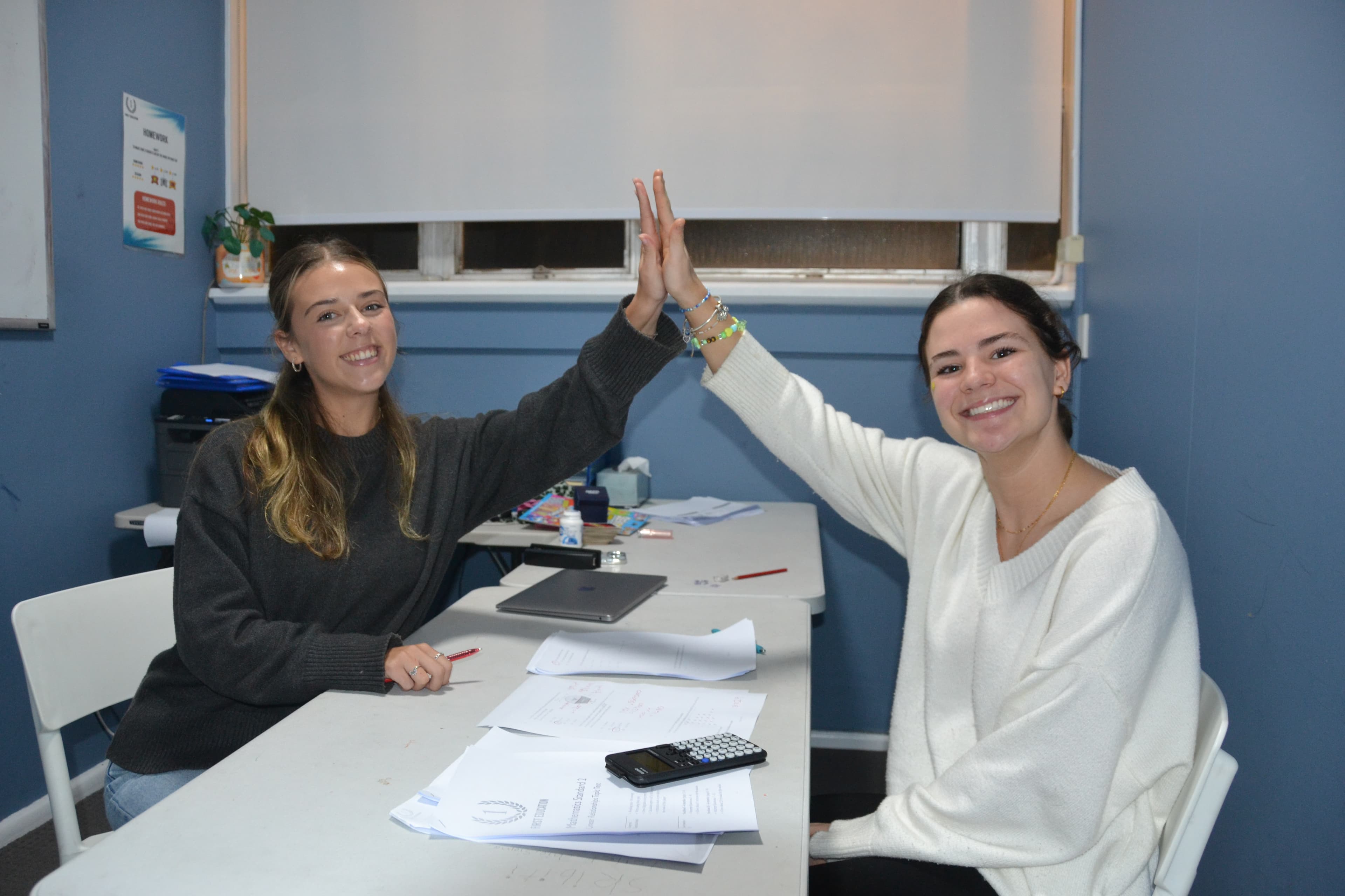 A First Education tutor and a Year 10 student high-fiving over completed maths papers at a centre desk.
