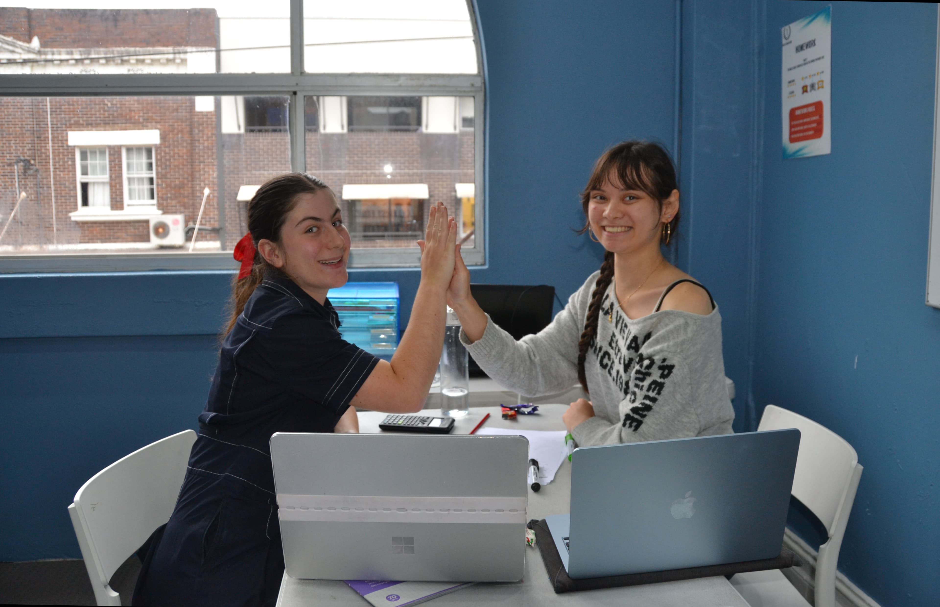 A First Education tutor and a Year 11 student giving a high five at a centre desk with open laptops and study materials.