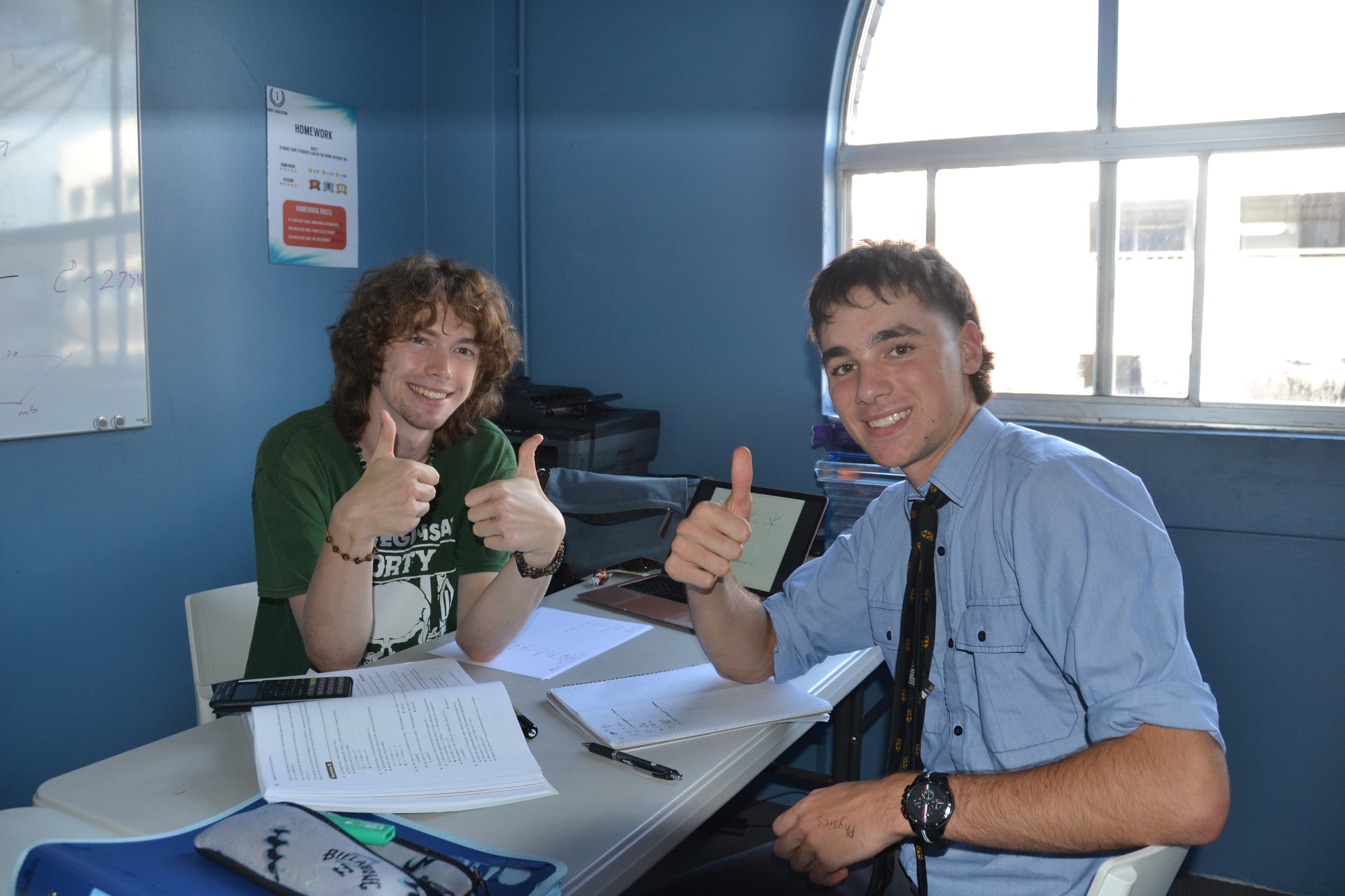 A First Education tutor and a Year 11 student giving double thumbs up at a centre desk with worksheets and study materials.