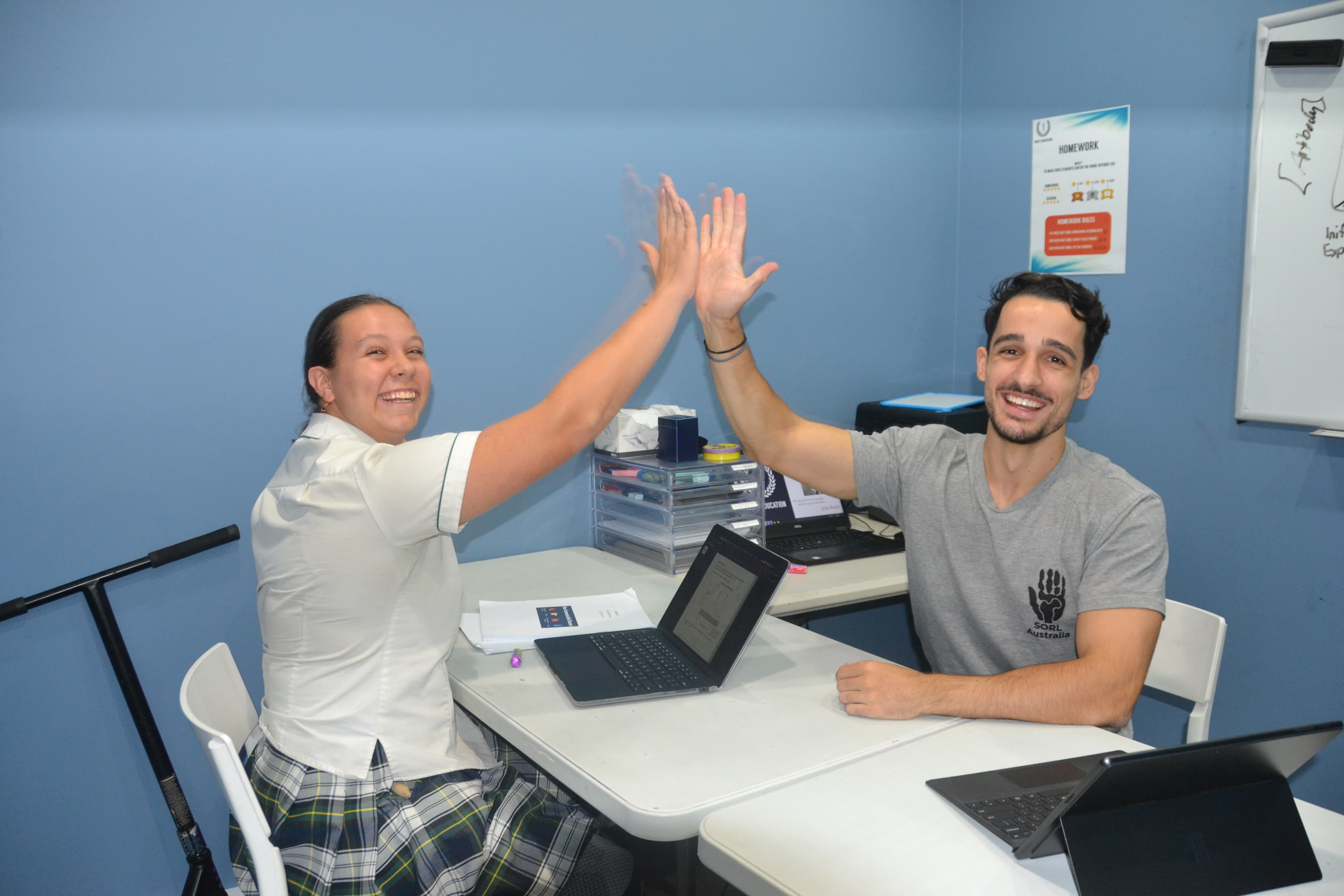 A First Education tutor and a Year 12 student sitting together at a centre desk with open worksheets and study materials.
