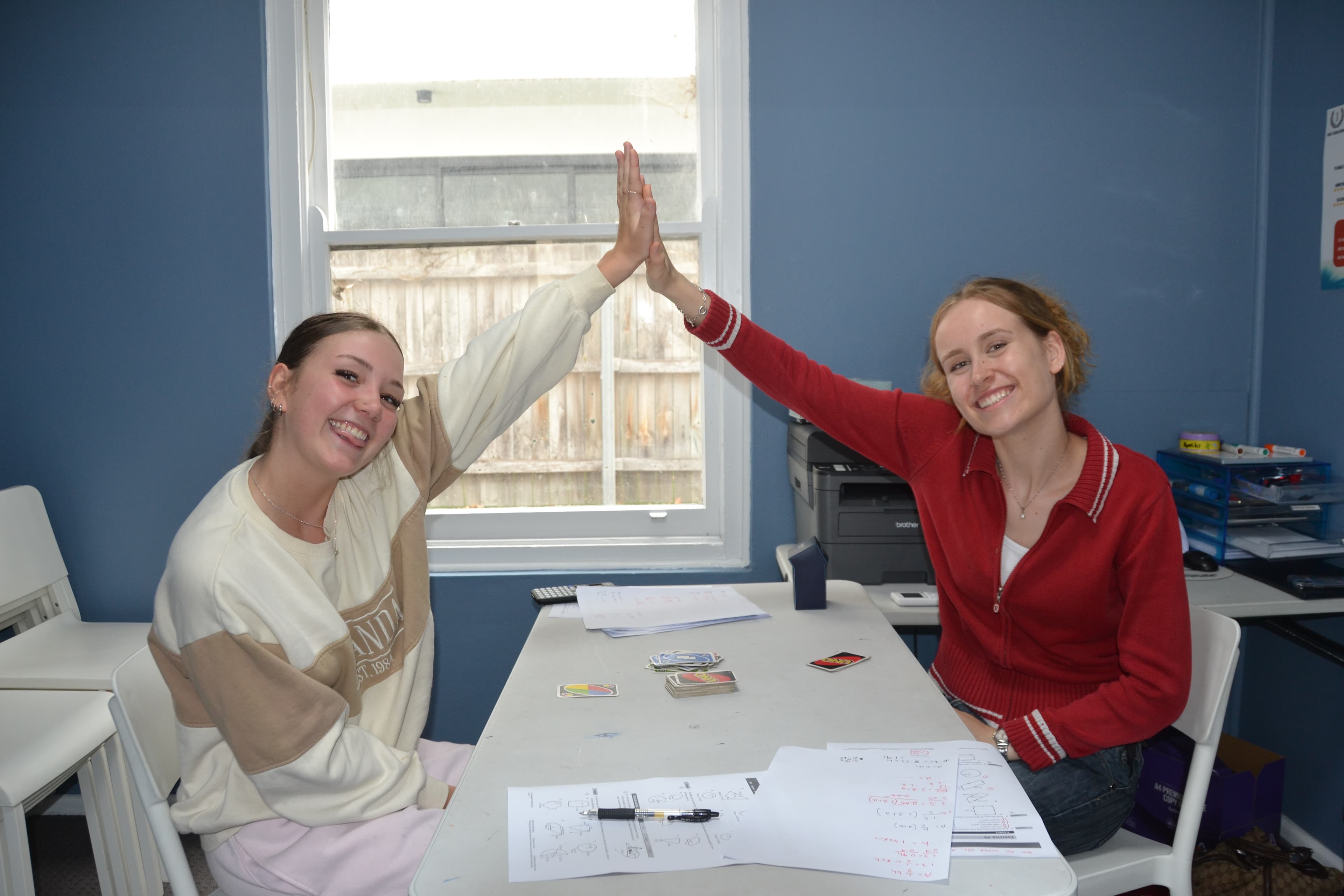 A First Education tutor and a student high-fiving at a centre desk with worksheets and playing cards.