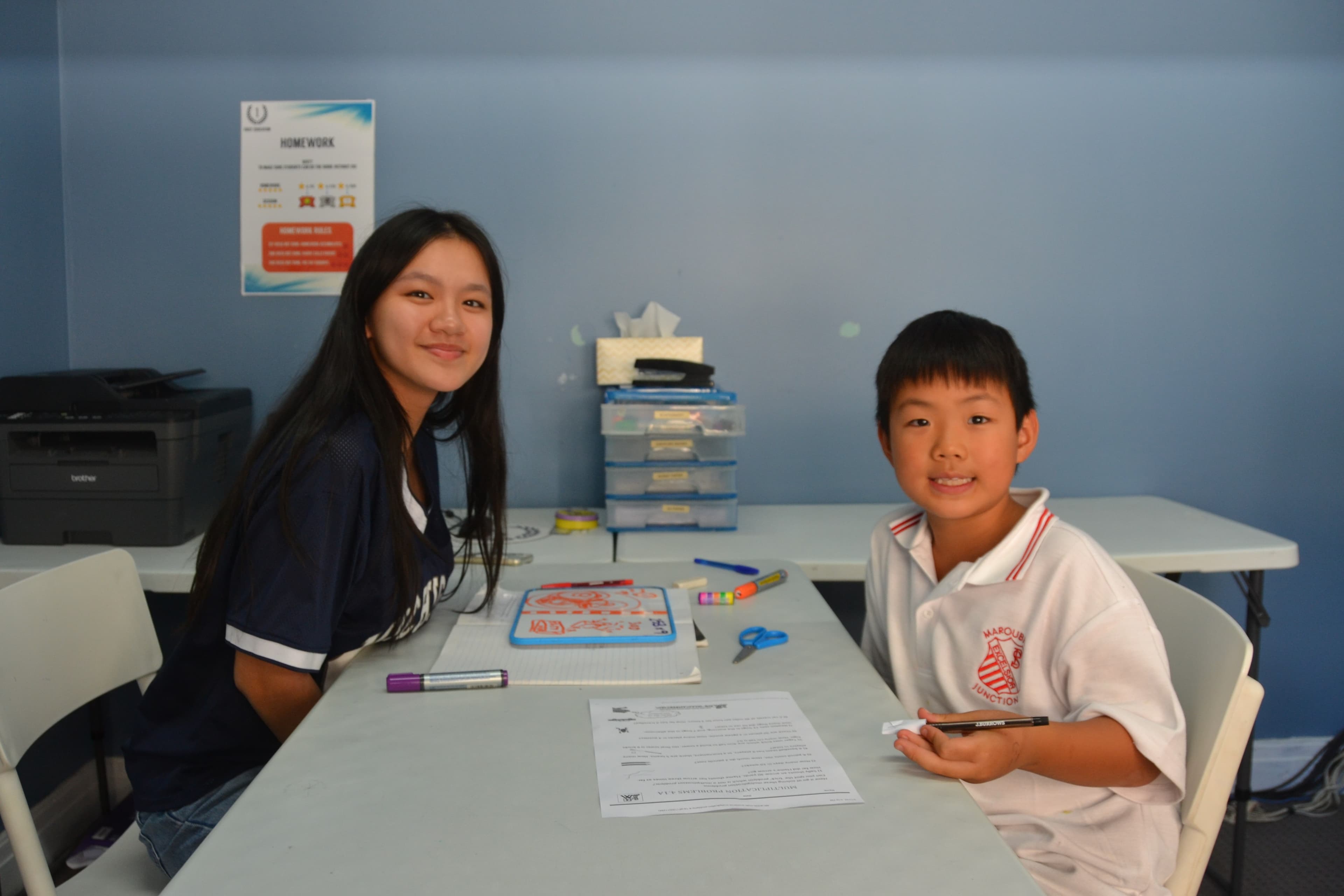 A First Education tutor and a Year 2 student in school uniform sitting together at a centre desk with colored markers and worksheets.