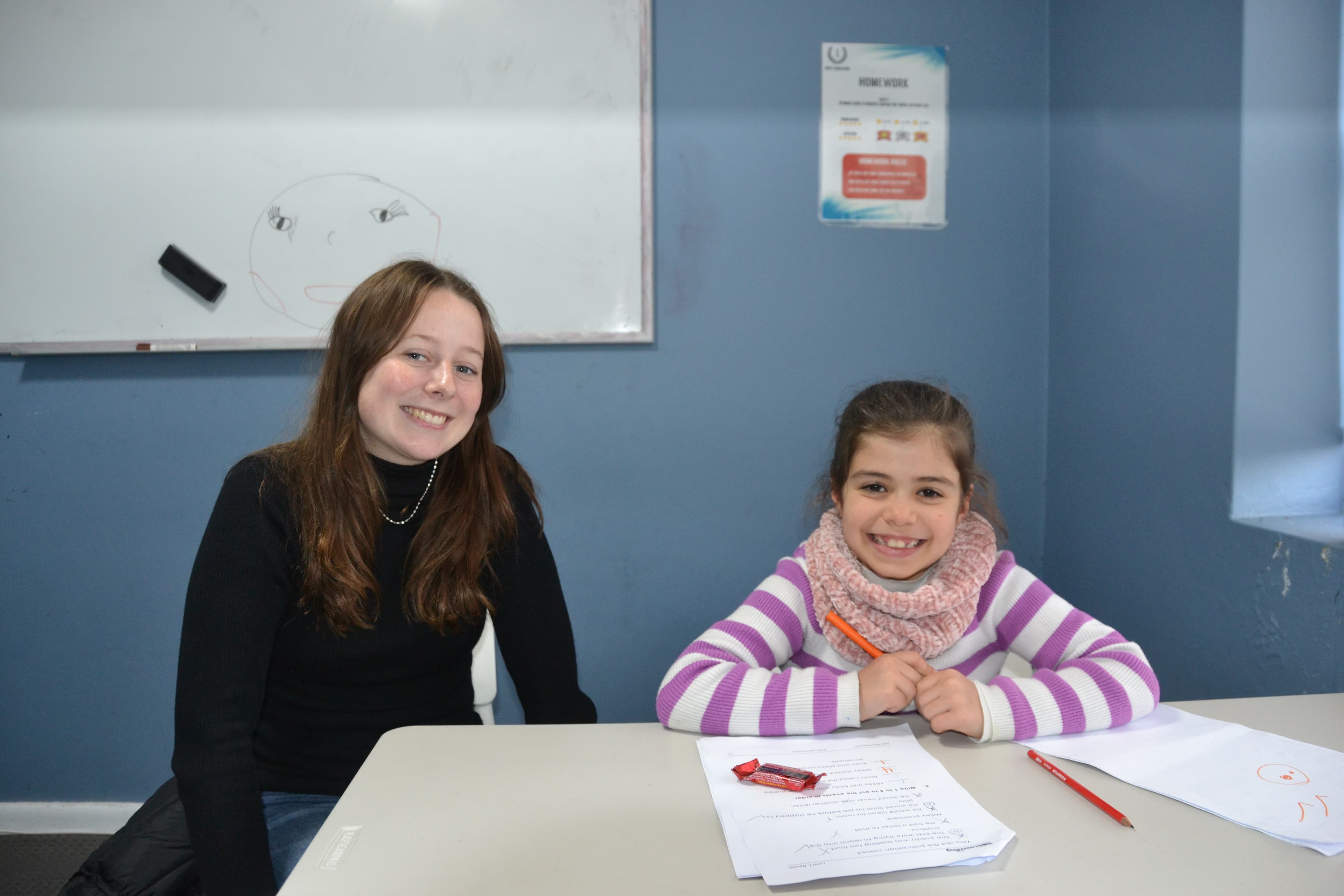 A First Education tutor and a Year 2 student sitting together at a centre desk with open worksheets and study materials.