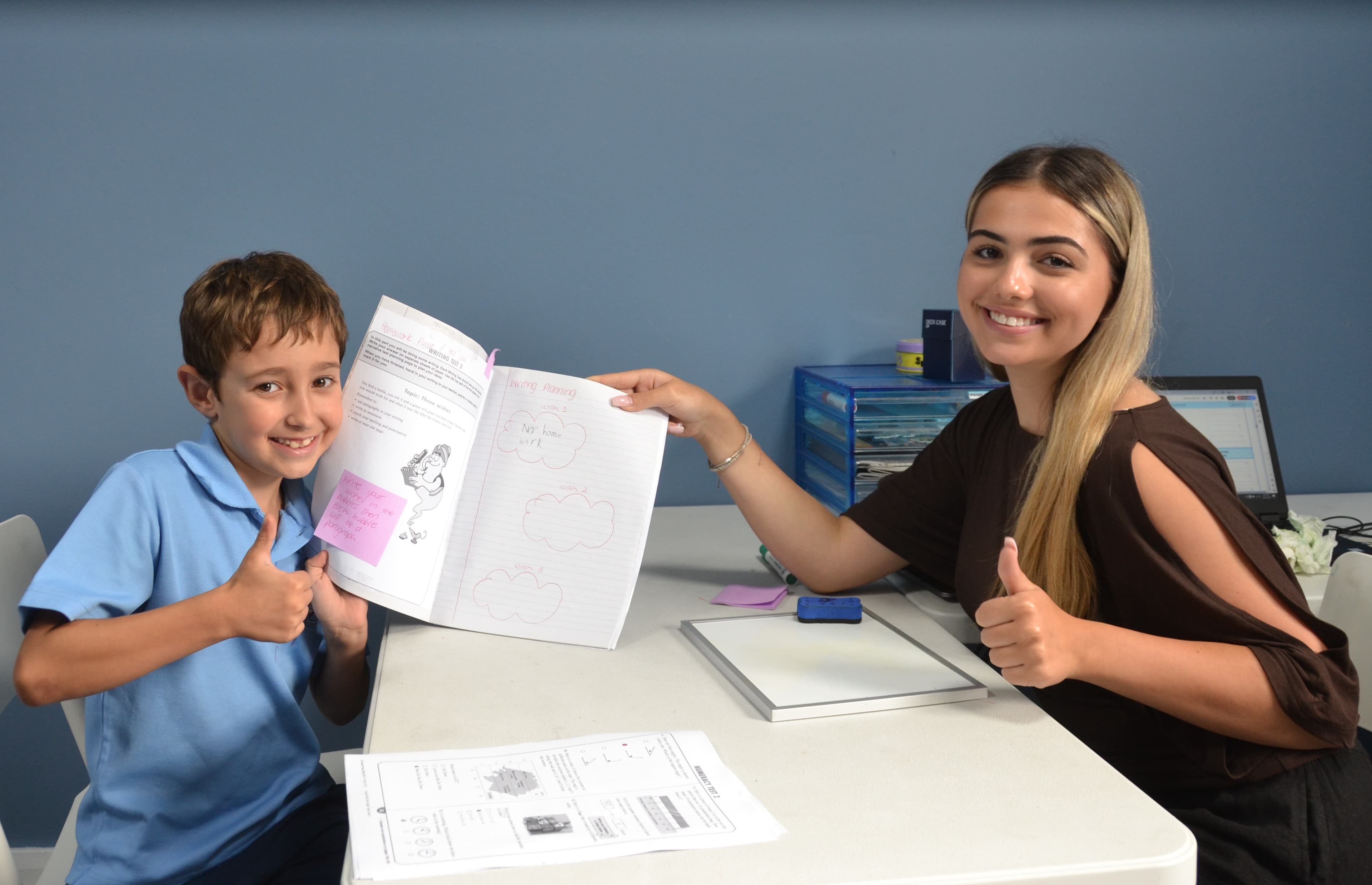 A First Education tutor and a Year 2 student giving a thumbs up while reviewing an open workbook at a centre desk.