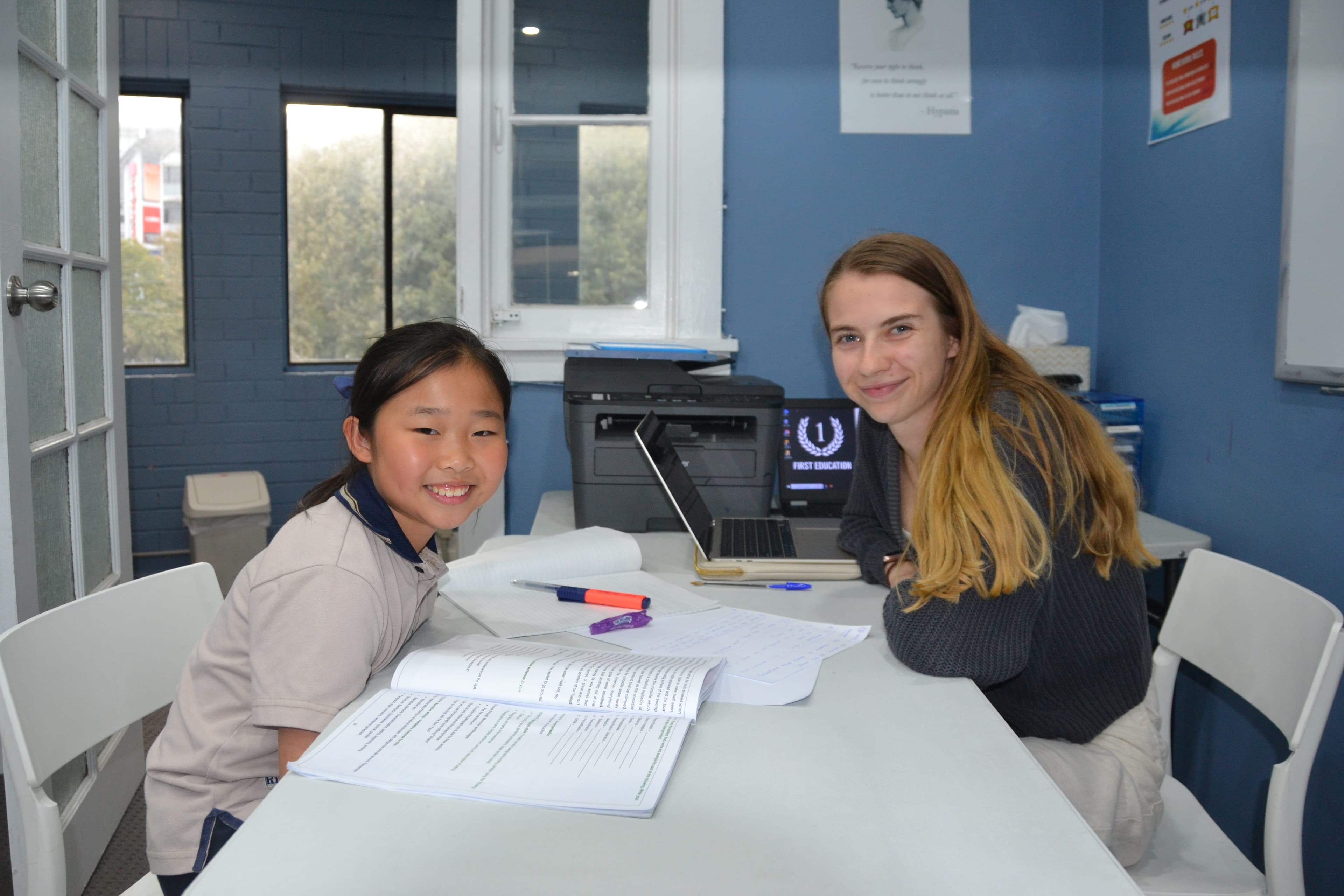 A First Education tutor and a Year 4 student sitting together at a centre desk with open worksheets and study materials.