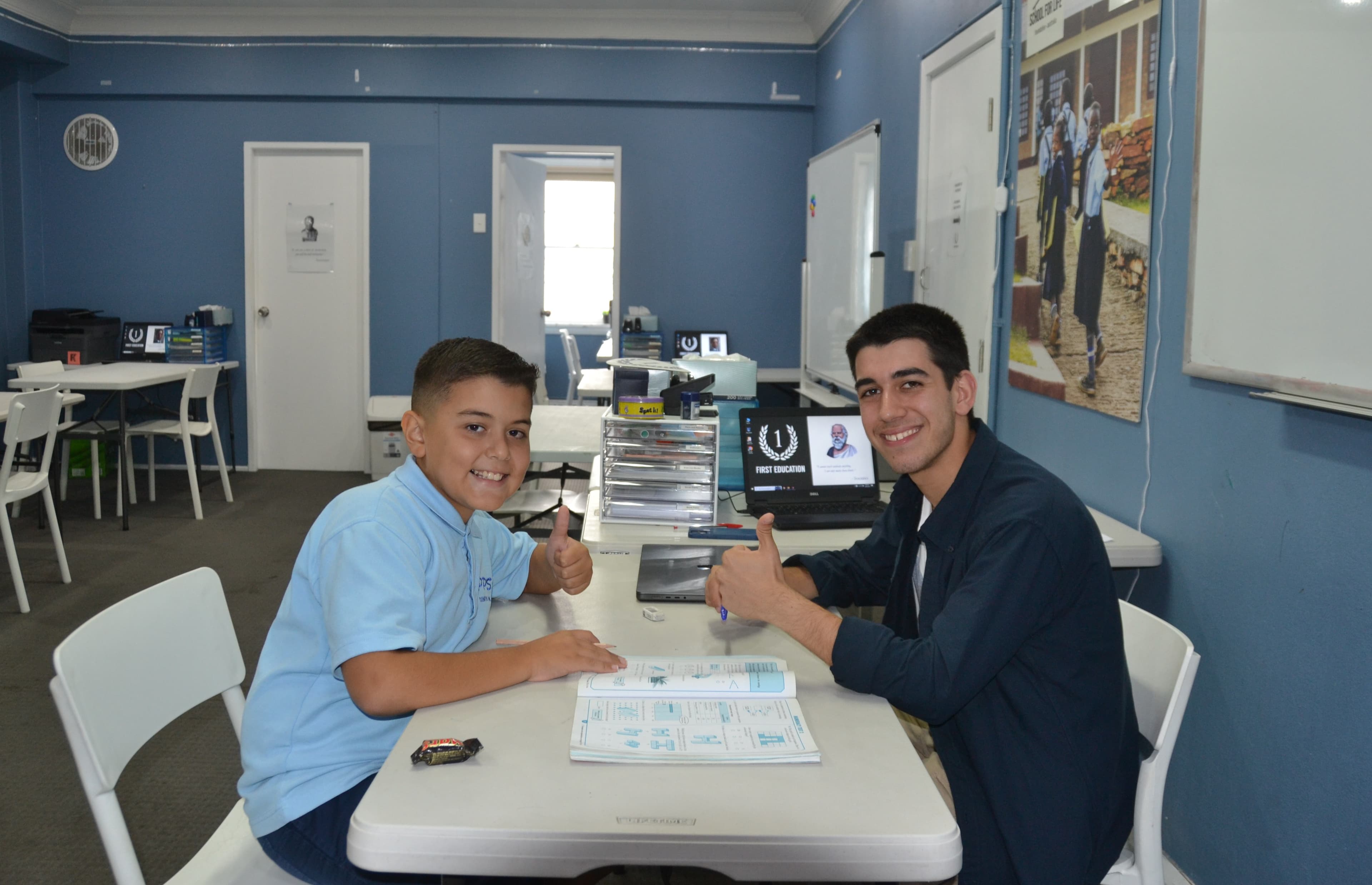 A First Education tutor and a Year 4 student in school uniform giving a thumbs up at a centre desk with worksheets and study materials.