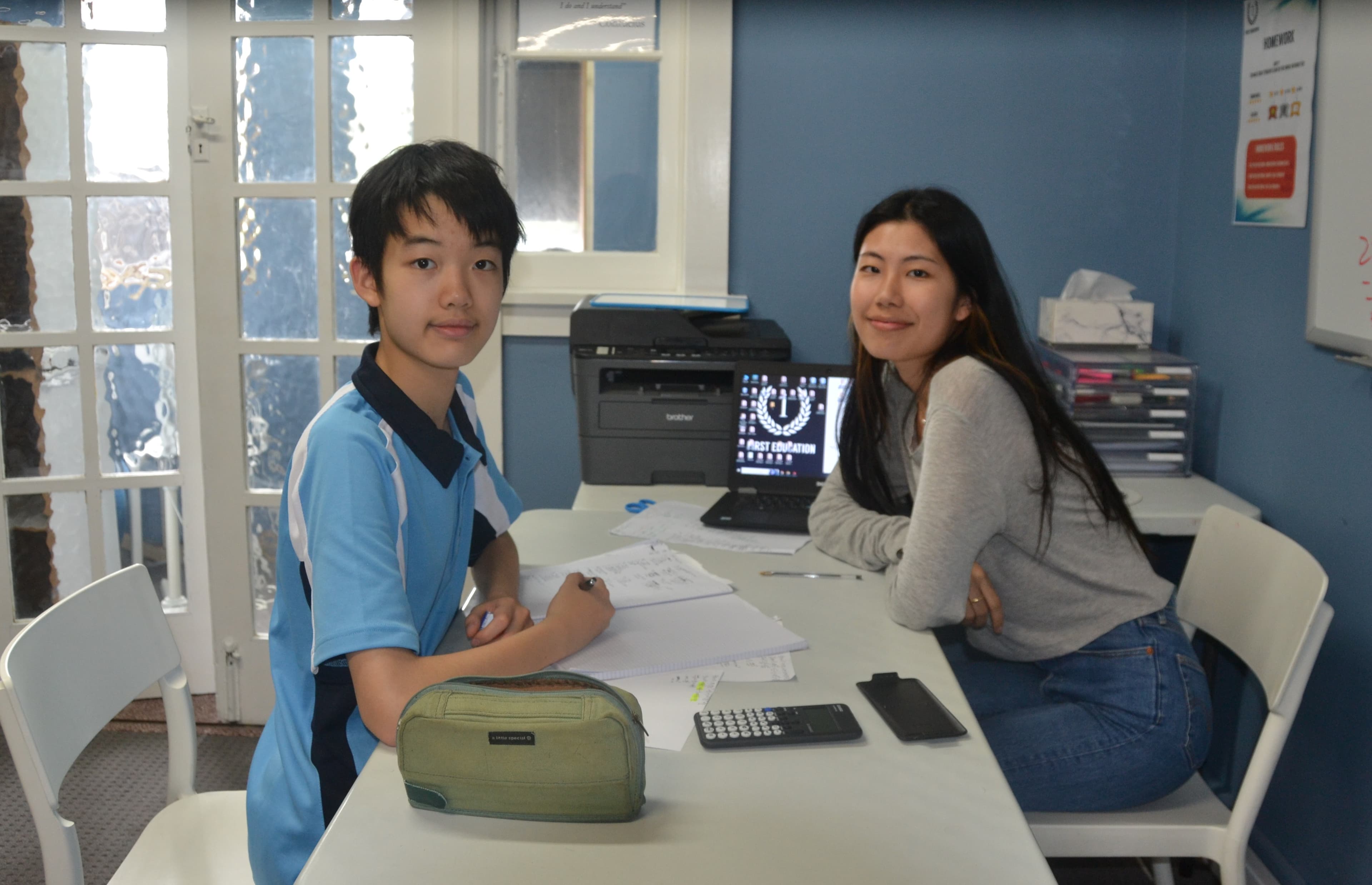 A First Education tutor and a Year 7 student sitting together at a centre desk with open worksheets and study materials.