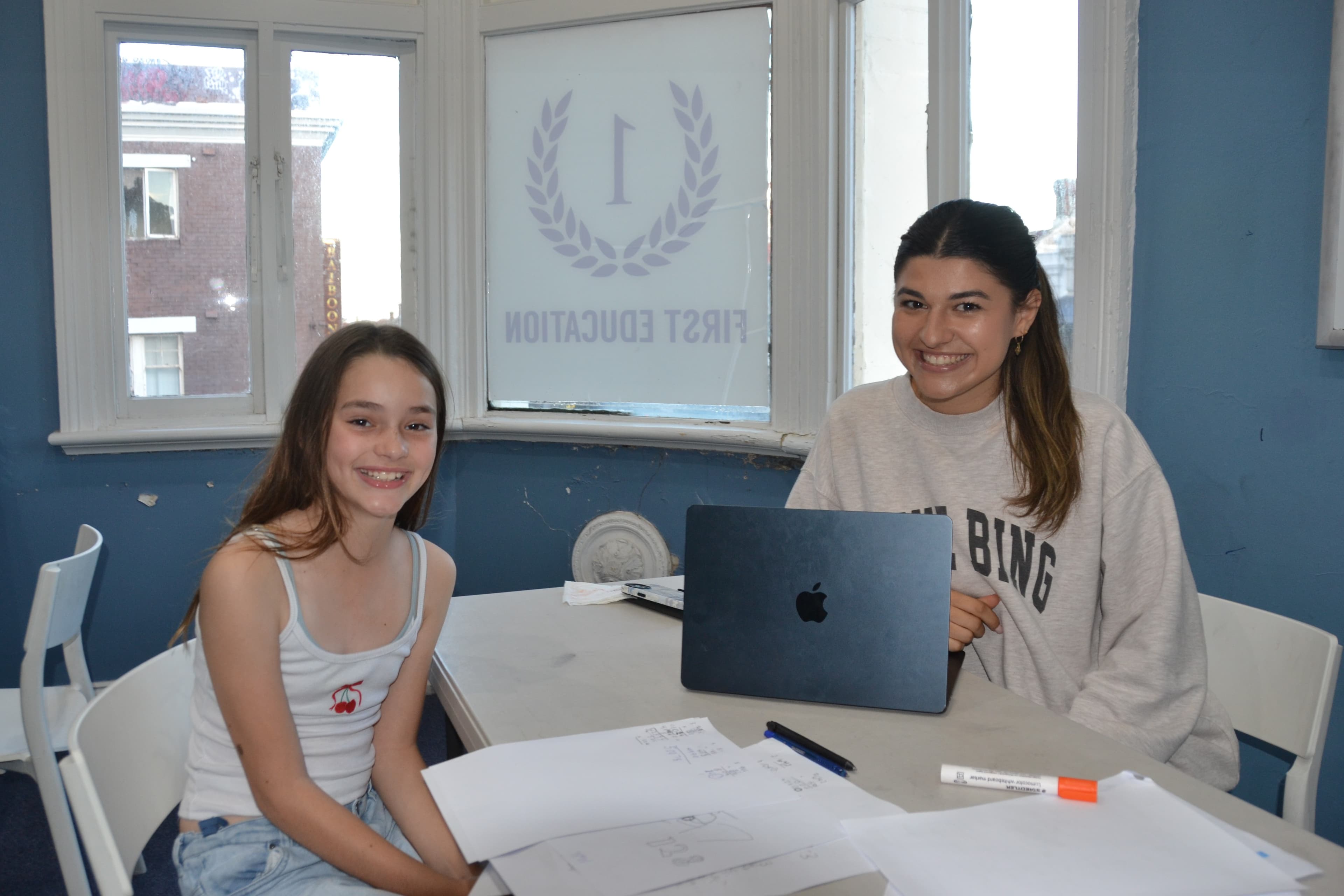 A First Education tutor and a Year 8 student sitting together at a centre desk with open workbooks and study materials.