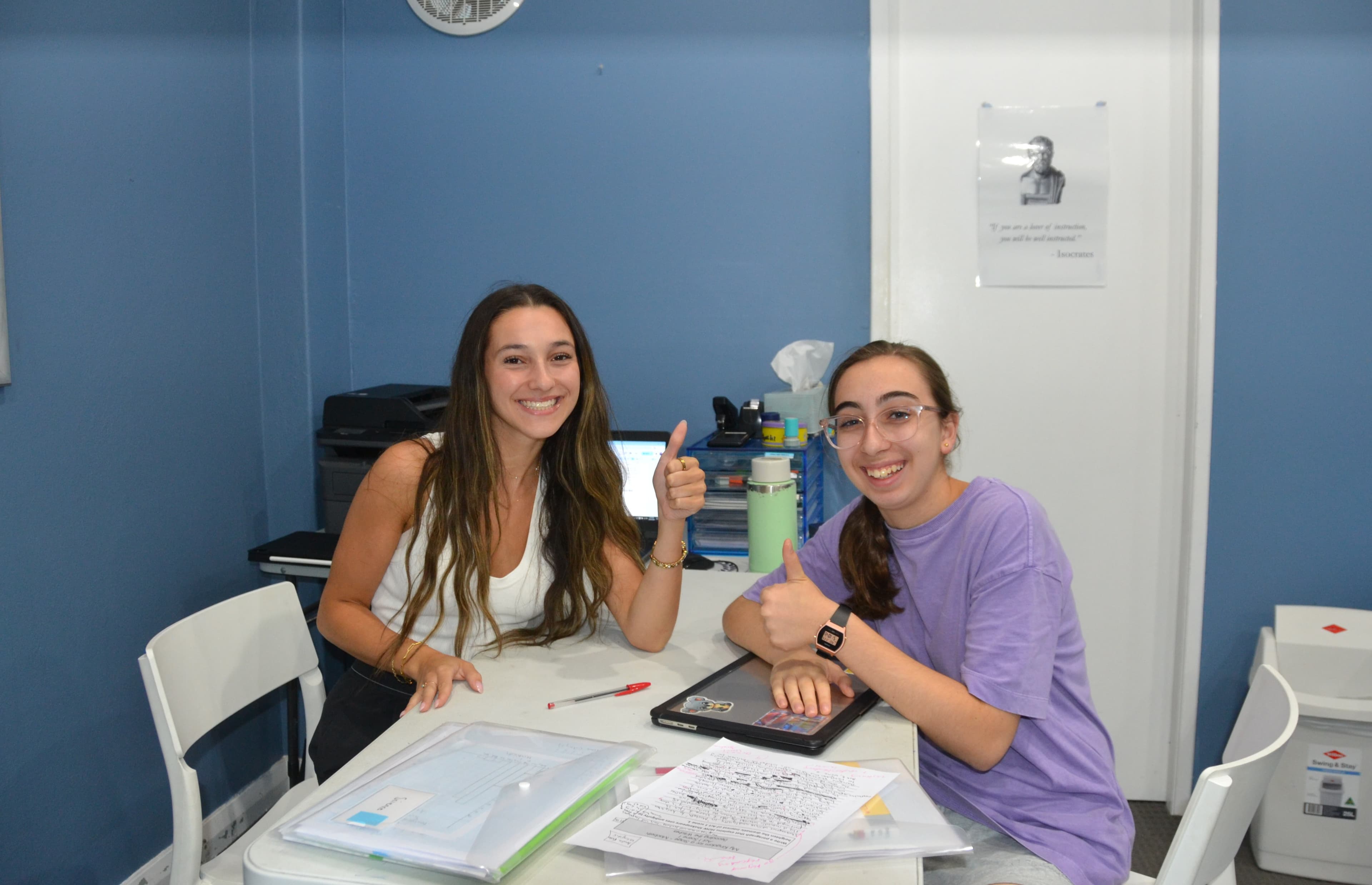 A First Education tutor and a student giving double thumbs up at a centre desk with study folders and an open laptop.