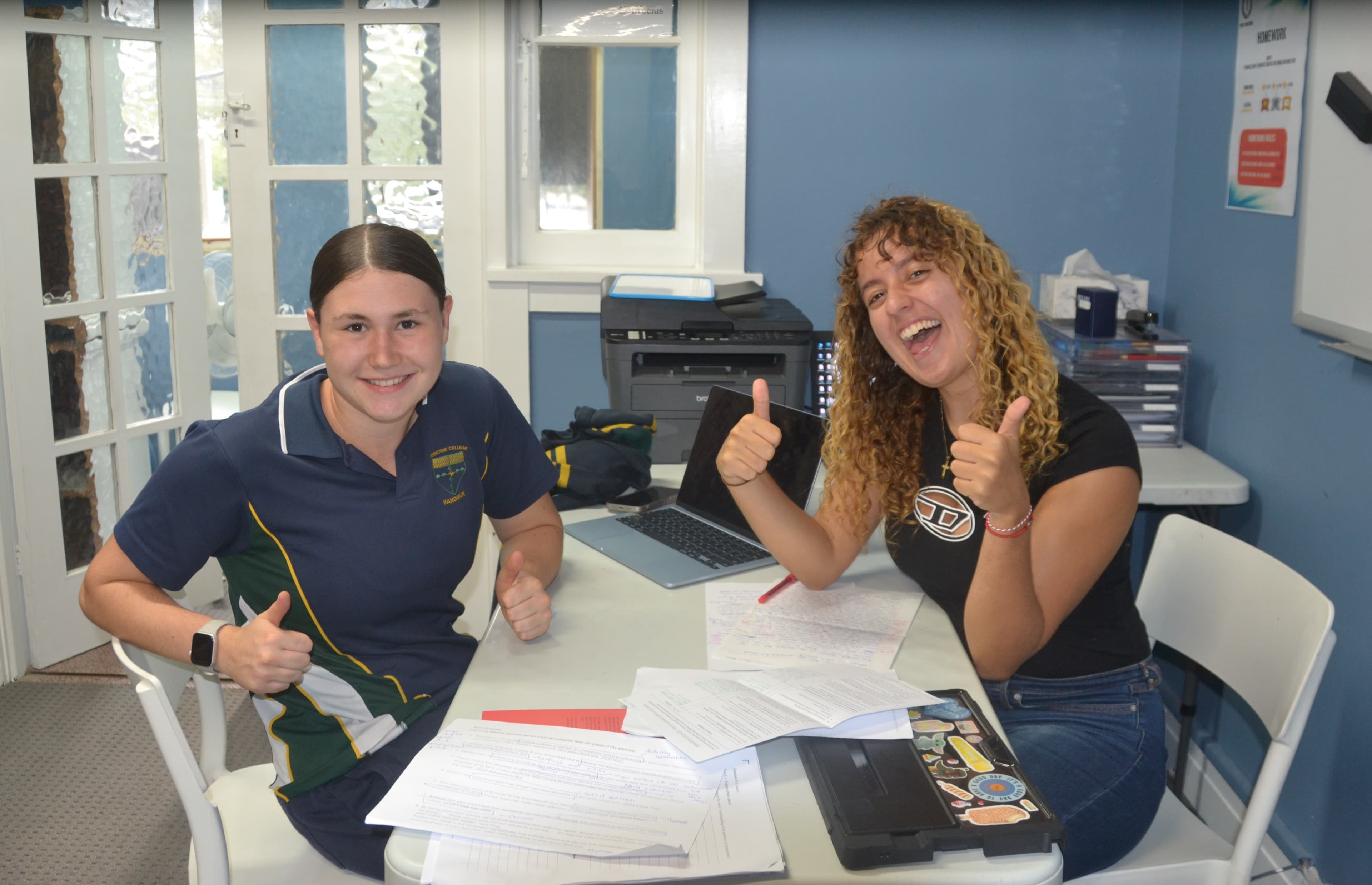 A First Education tutor and a student in school uniform laughing and giving double thumbs up at a centre desk with an open laptop.