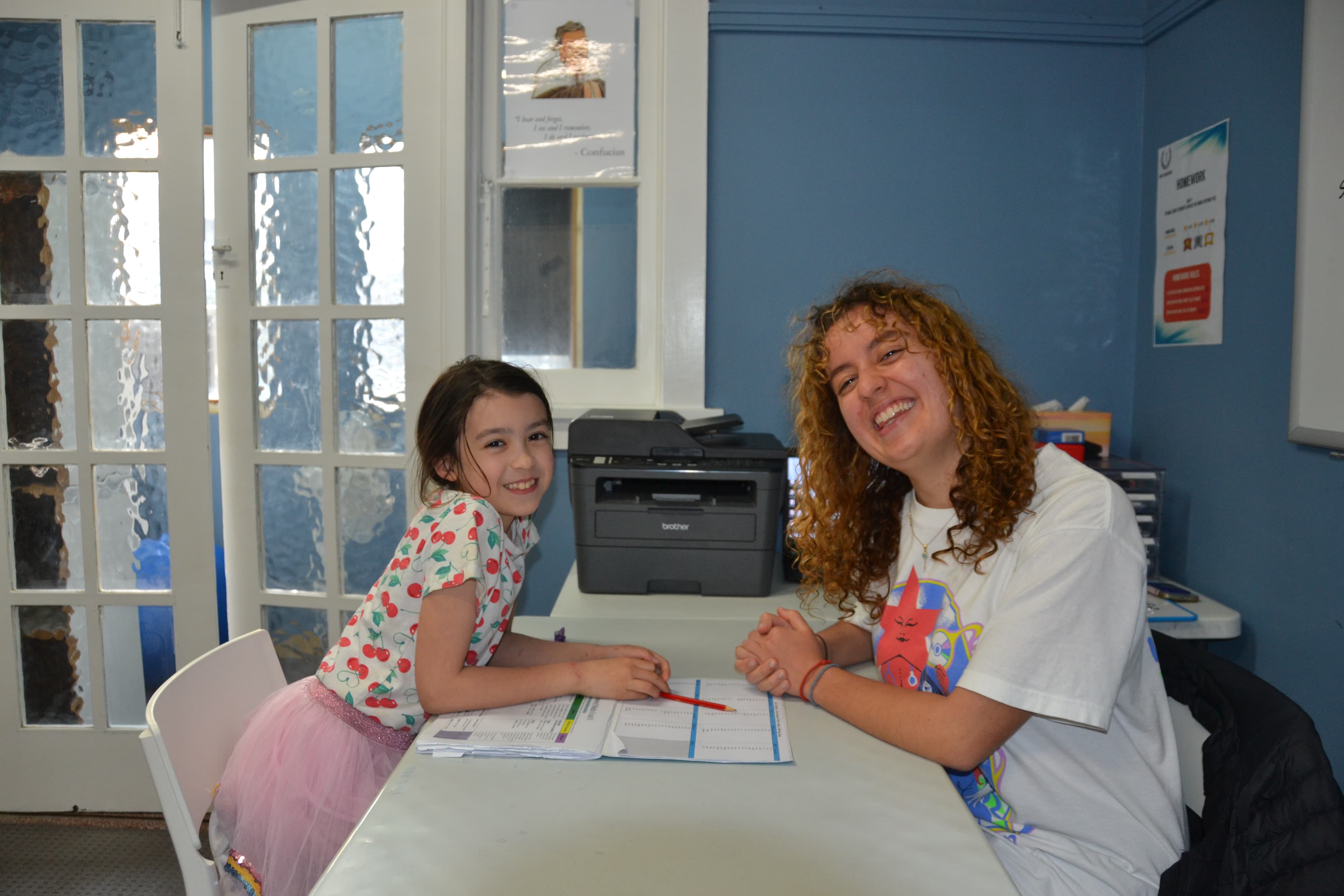 A First Education tutor and a student sitting together at a centre desk with worksheets and study materials.