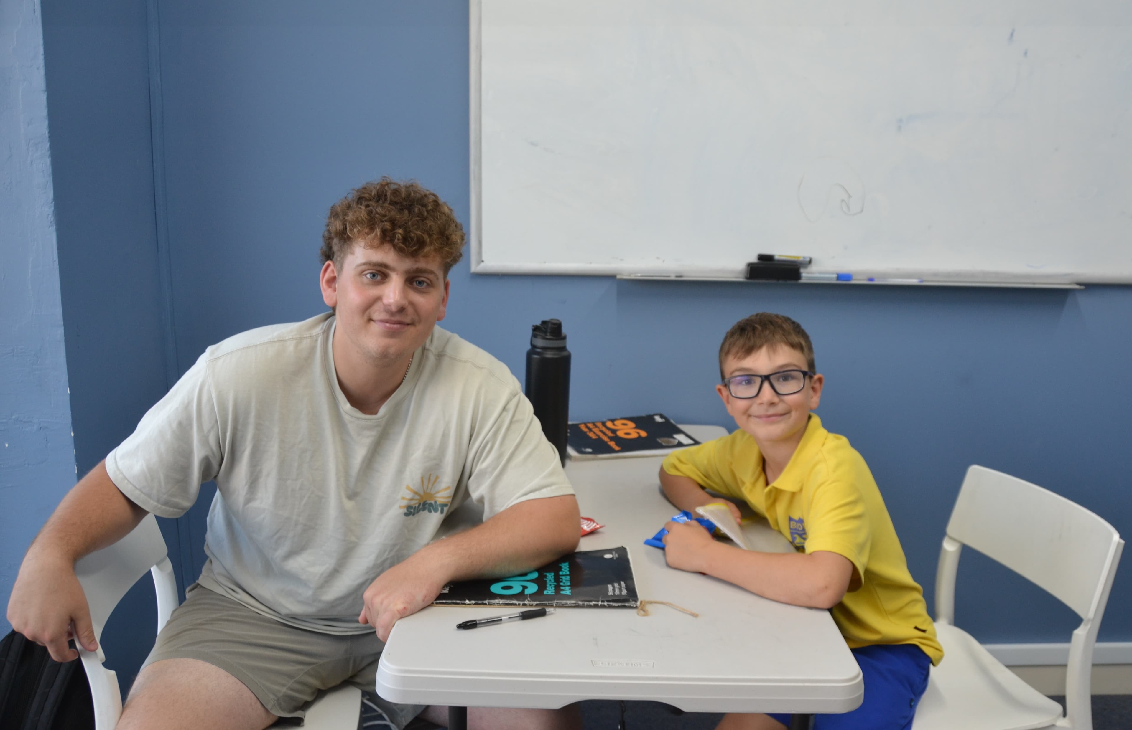 A First Education tutor and a primary school student in school uniform sitting together at a centre desk with exercise books, near a whiteboard.