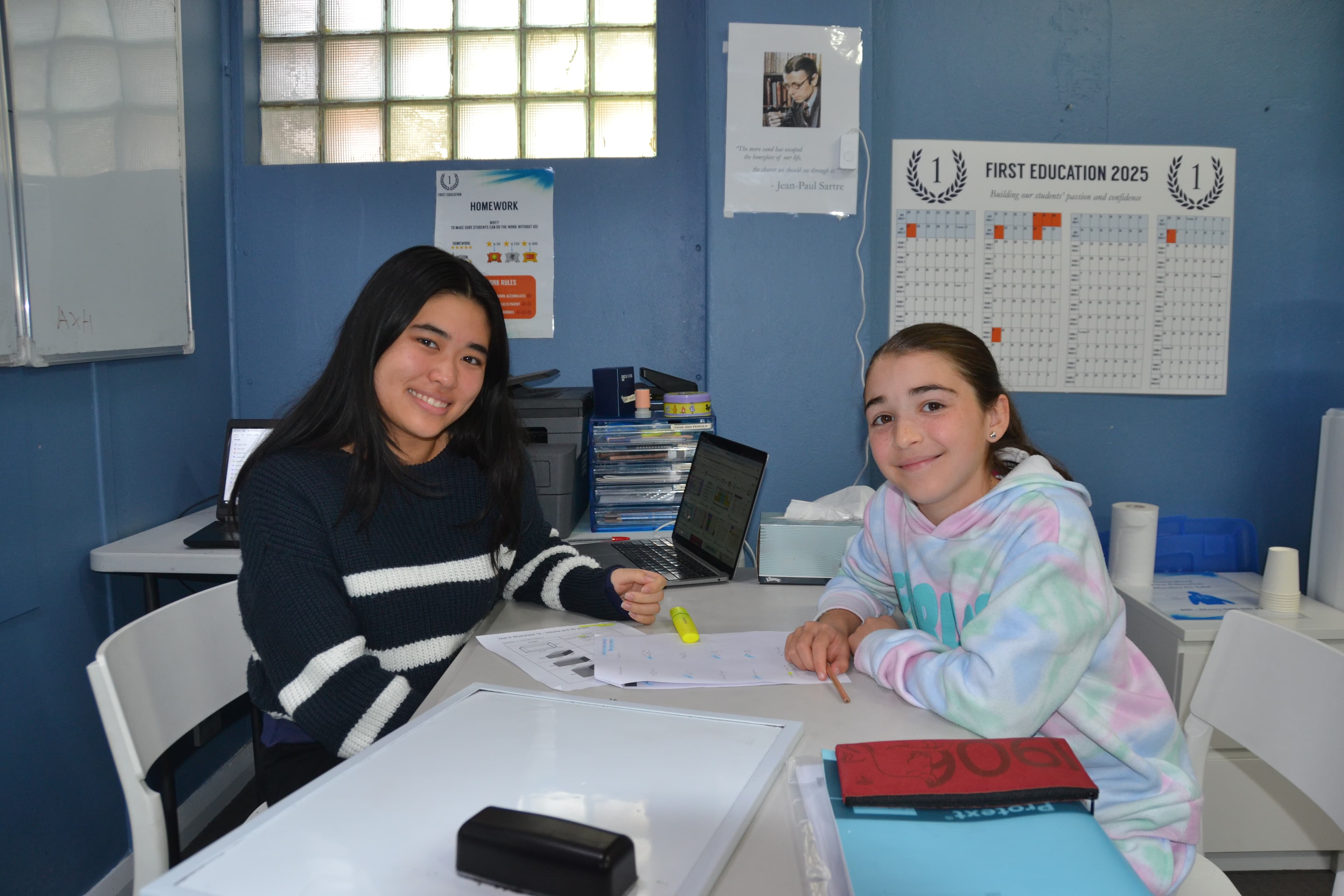 A First Education tutor and a Year 5 student sitting together at a centre desk with open worksheets and markers.