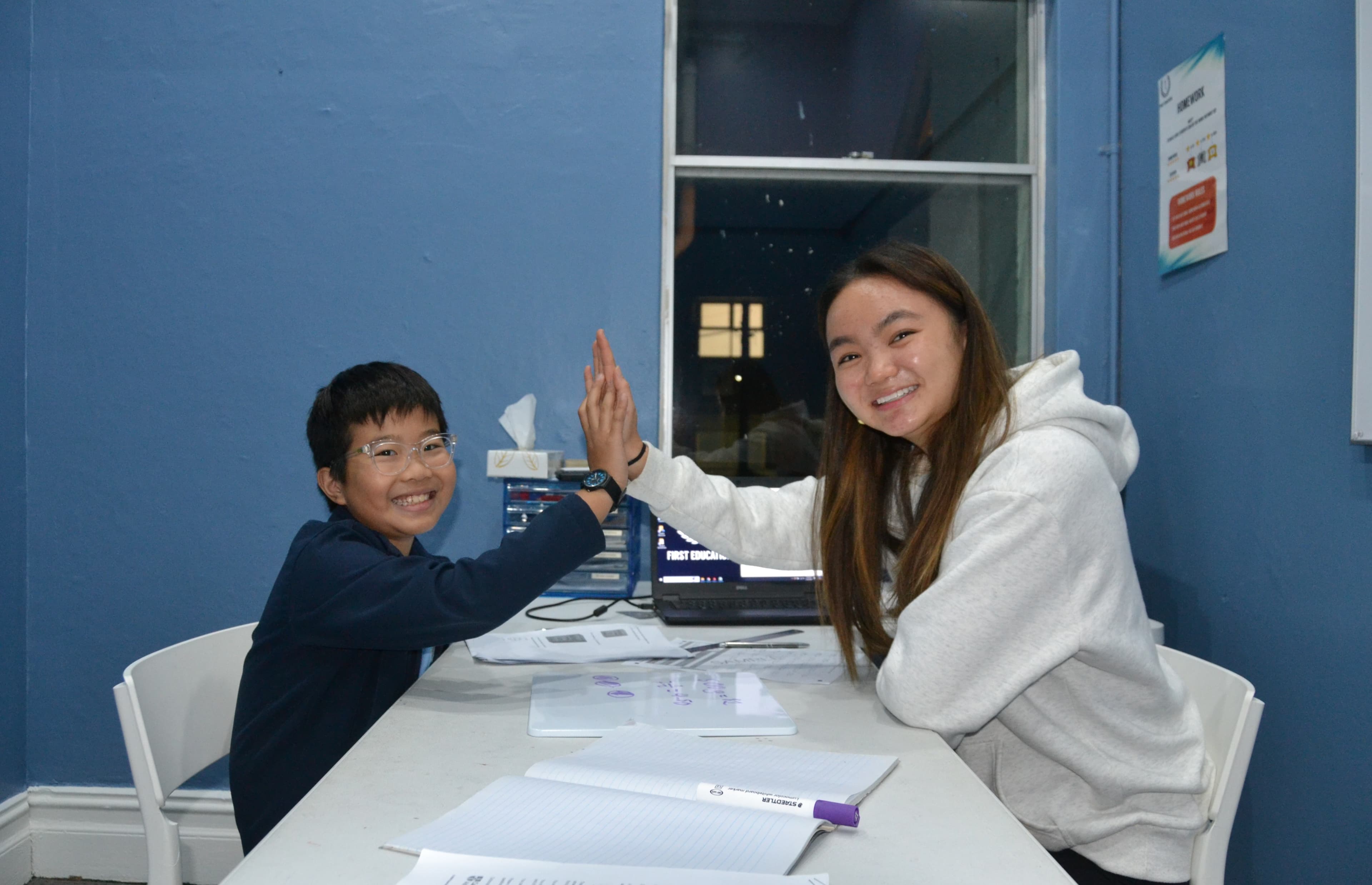 A First Education tutor and a Year 5 student sitting together at a centre desk with open workbooks and a laptop.